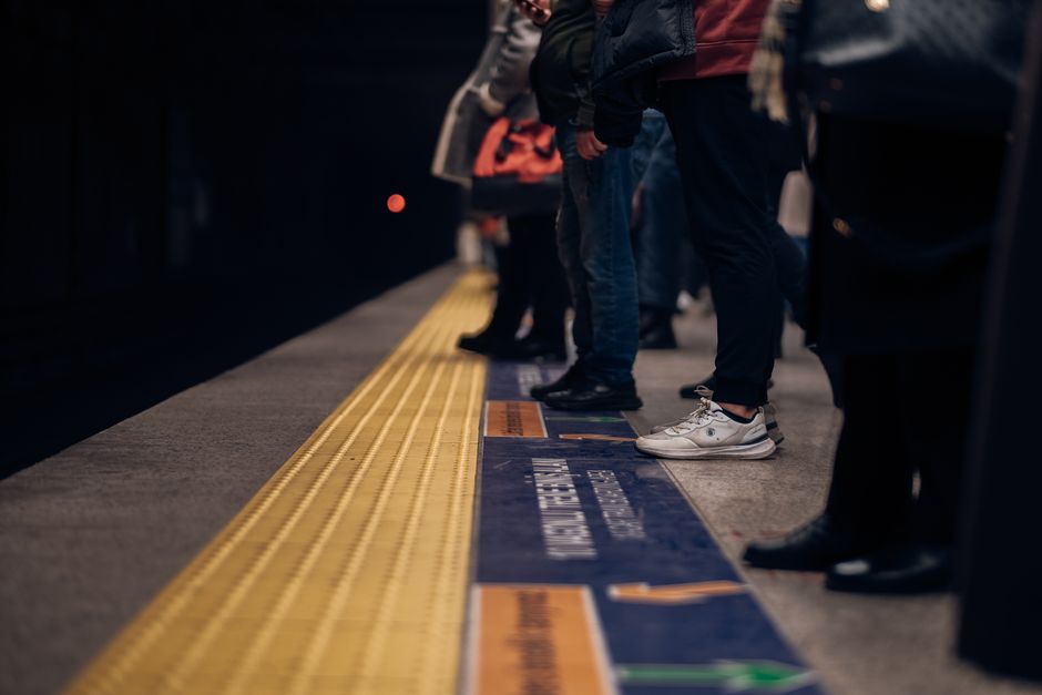 Photograph of a modern subway station platform featuring a sleek, reflective black ceiling and bright overhead lighting. The platform surface is tiled in light grey with a yellow tactile paving strip 